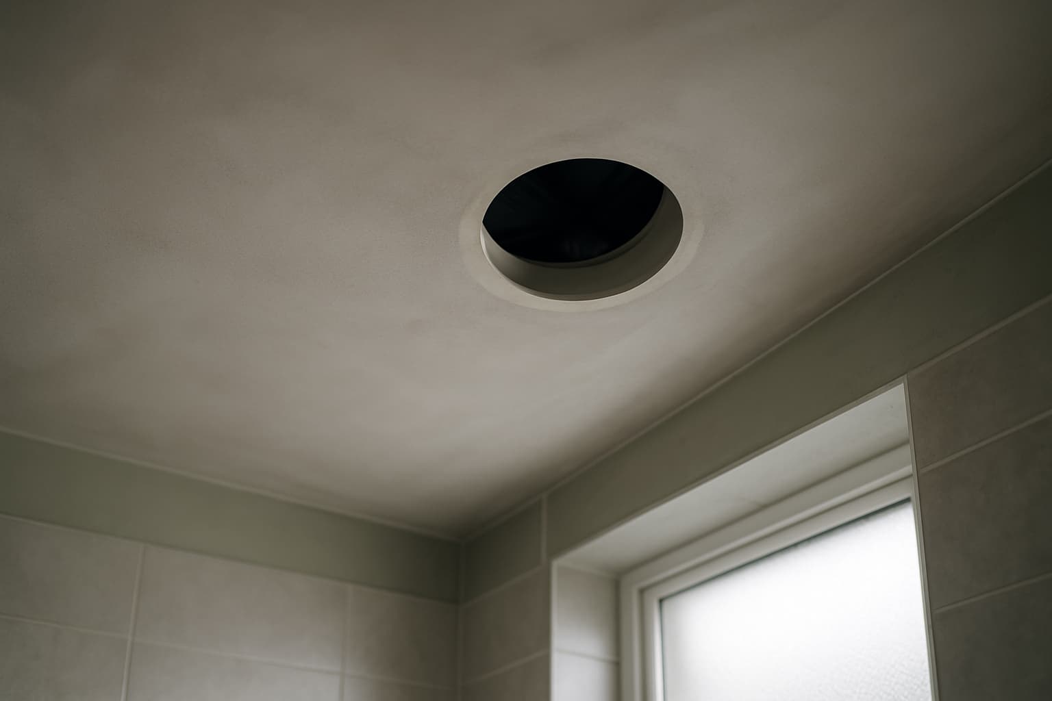 Wit gestuukt badkamerplafond met ronde uitsparing voor een afzuigventiel, met zichtbare ventilatiebuis erboven en daglicht uit een matglazen raam.